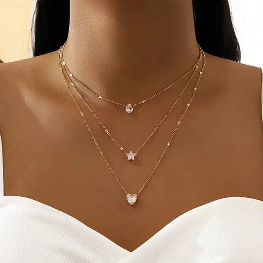 A close-up of a model's neck and chest wearing a three-layered rose gold necklace set with clear crystal pendants shaped as a heart, a star, and a small round stone.